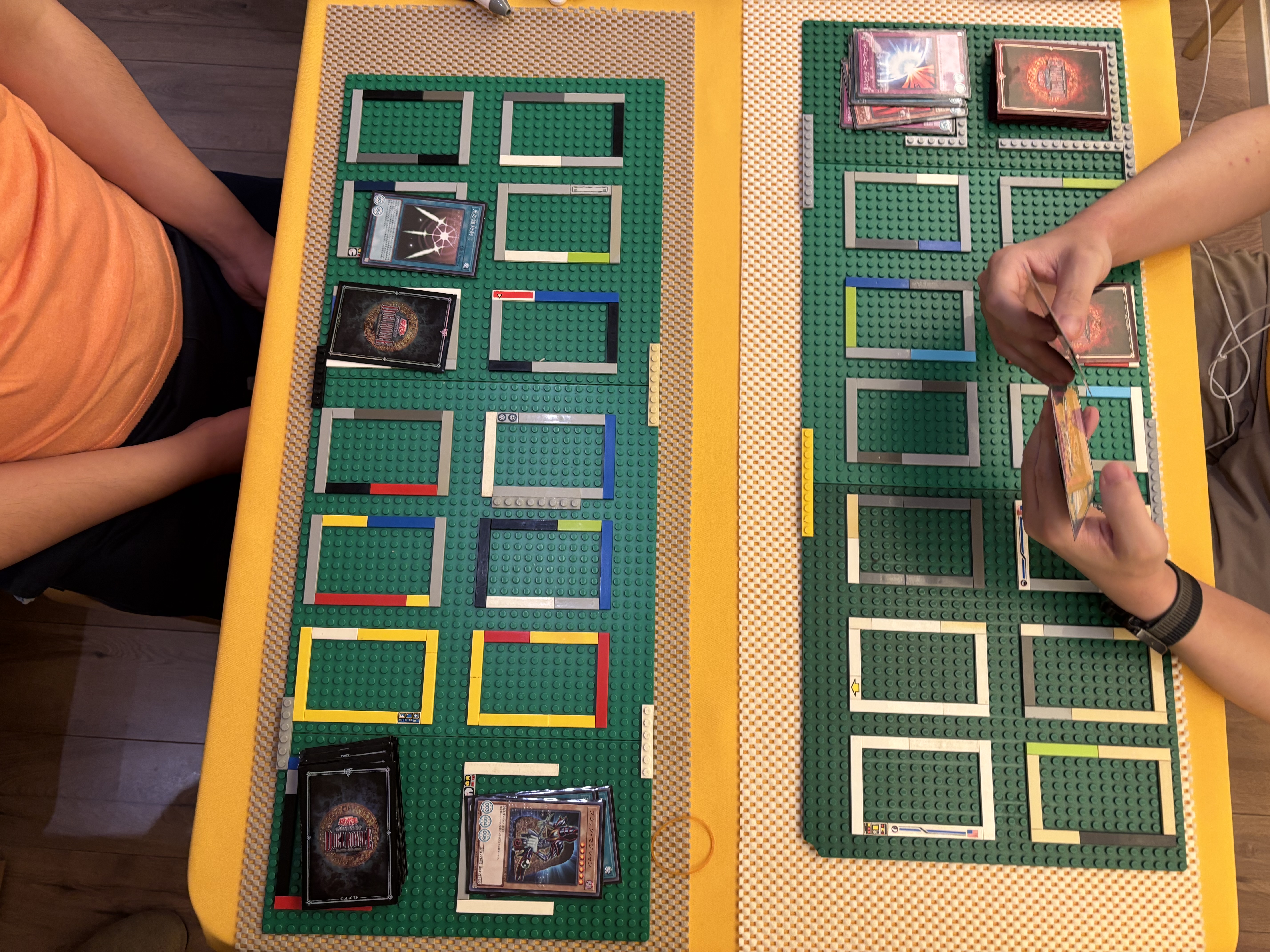 Playing the accessible edition Yu-Gi-Oh! The play mat is made of Lego so cards don't go outside the frame. The left player has an attack position monster, face-down defense monster on the field, and one spell card activated. The right player's field has no cards.